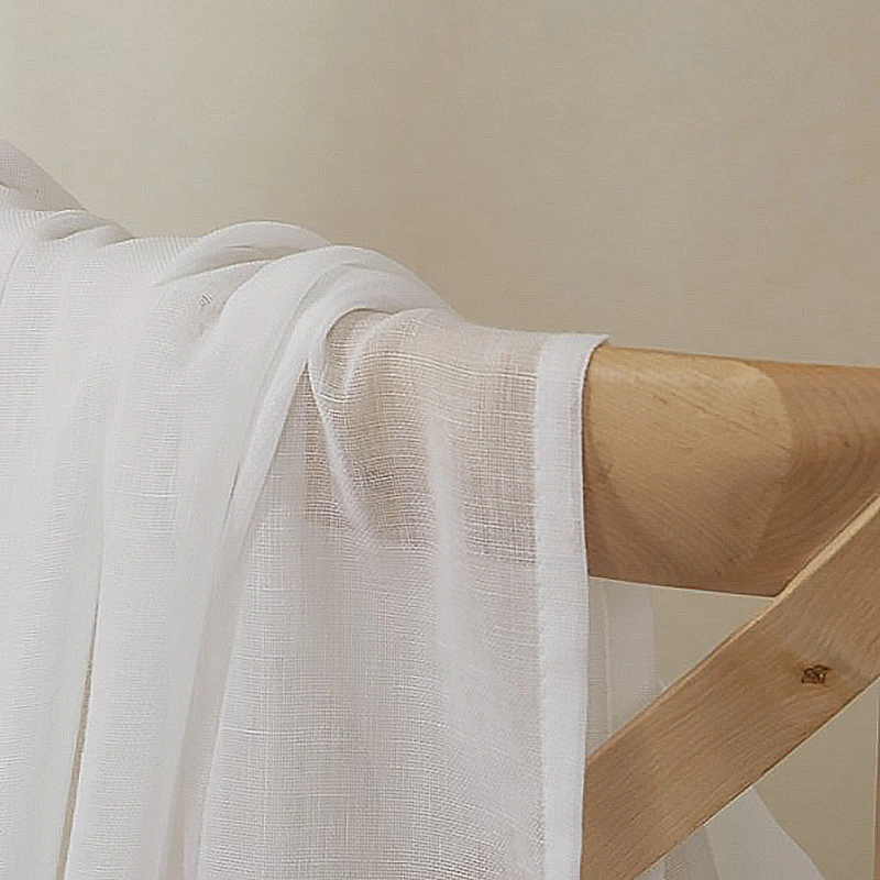 A close-up of the crisp white fabric of semi-sheer linen curtains and the smooth wooden curtain rod.